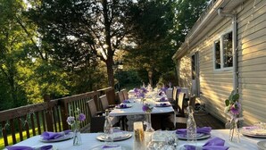 An outdoor dining area with tables set for a meal, surrounded by trees and a wooden deck.