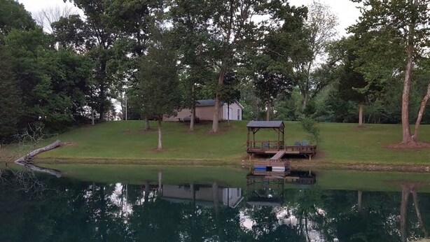 A tranquil pond with a wooden dock and a gazebo, surrounded by lush greenery and a house in the background.