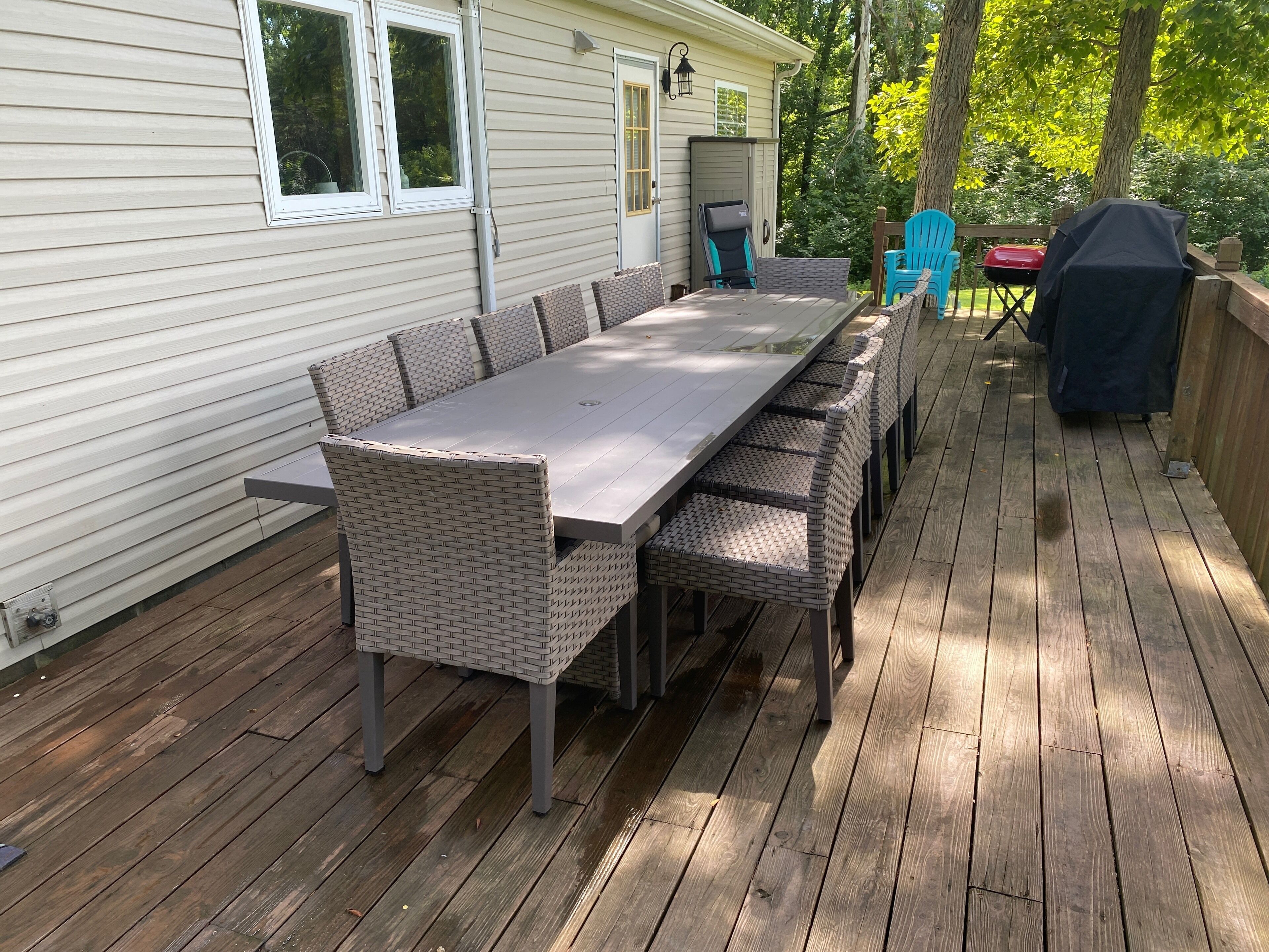 A covered deck with a long dining table and chairs, a grill, and a chair with a blue cushion.