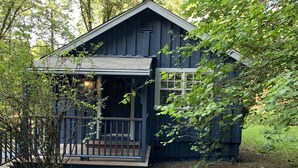 Eine kleine, dunkle Holzhütte mit Veranda und Fenster, umgeben von Bäumen und Grün.