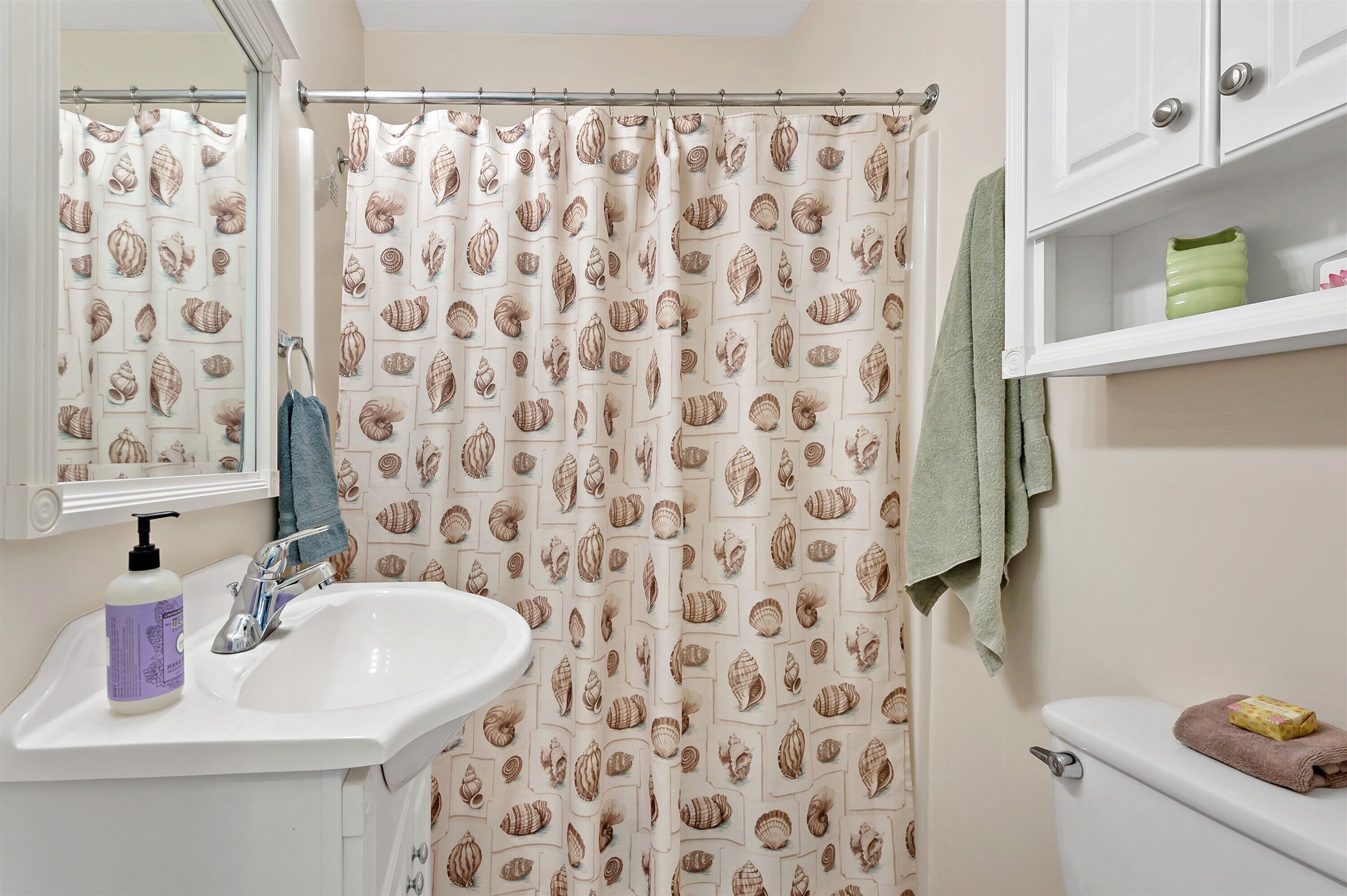 Une salle de bain avec un rideau de douche orné d’un motif de coquillages, un lavabo avec un distributeur de savon et des toilettes sur lesquelles est posée une serviette.