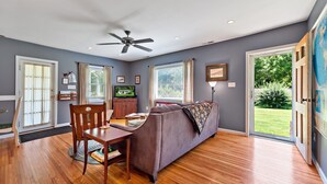 A spacious living room with a ceiling fan, wooden flooring, a gray sofa, a dining table with chairs, and a television. - Stay and Play @ Railway Rest, near AVL, trails, falls (Old Fort)