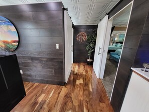 A hallway with dark wooden walls, a wooden floor, and a door leading to a bedroom with a blue bed.