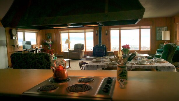 A cozy living room with a large window, a fireplace, and a kitchen area with a stove. - Historic - Private 28 Acres - 180° Ocean View! - Carolyn's suite (Homer)