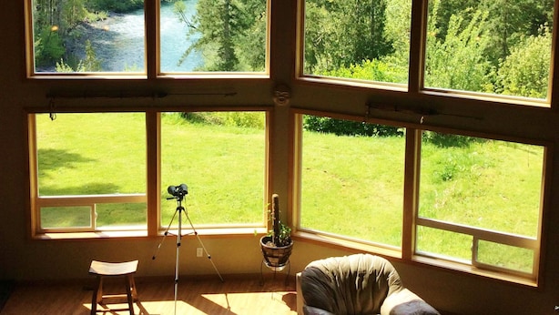 Ein Zimmer mit großen Fenstern, das einen Blick auf einen Fluss und ein Waldgebiet bietet. Ein Stativ mit einer Kamera, ein Stuhl und ein kleiner Hocker sind vorhanden.