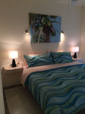 A bedroom with a bed, two bedside lamps, a painting of a palm tree, and a patterned bedspread. - Quiet and serene ocean front 2-bedroom 2-bathroom condo, steps from the sand! (Puerto Morelos)