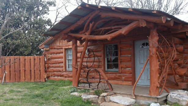 A rustic log cabin with a porch supported by tree branches, a wooden fence, and a stone pathway leading to the door.