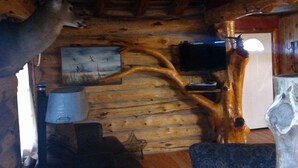 A rustic cabin interior with wooden walls, a mounted deer head, a mounted elk head, and a painting of a landscape.