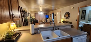A kitchen with a double sink, wooden cabinets, a countertop with a lamp, and a television in the background.