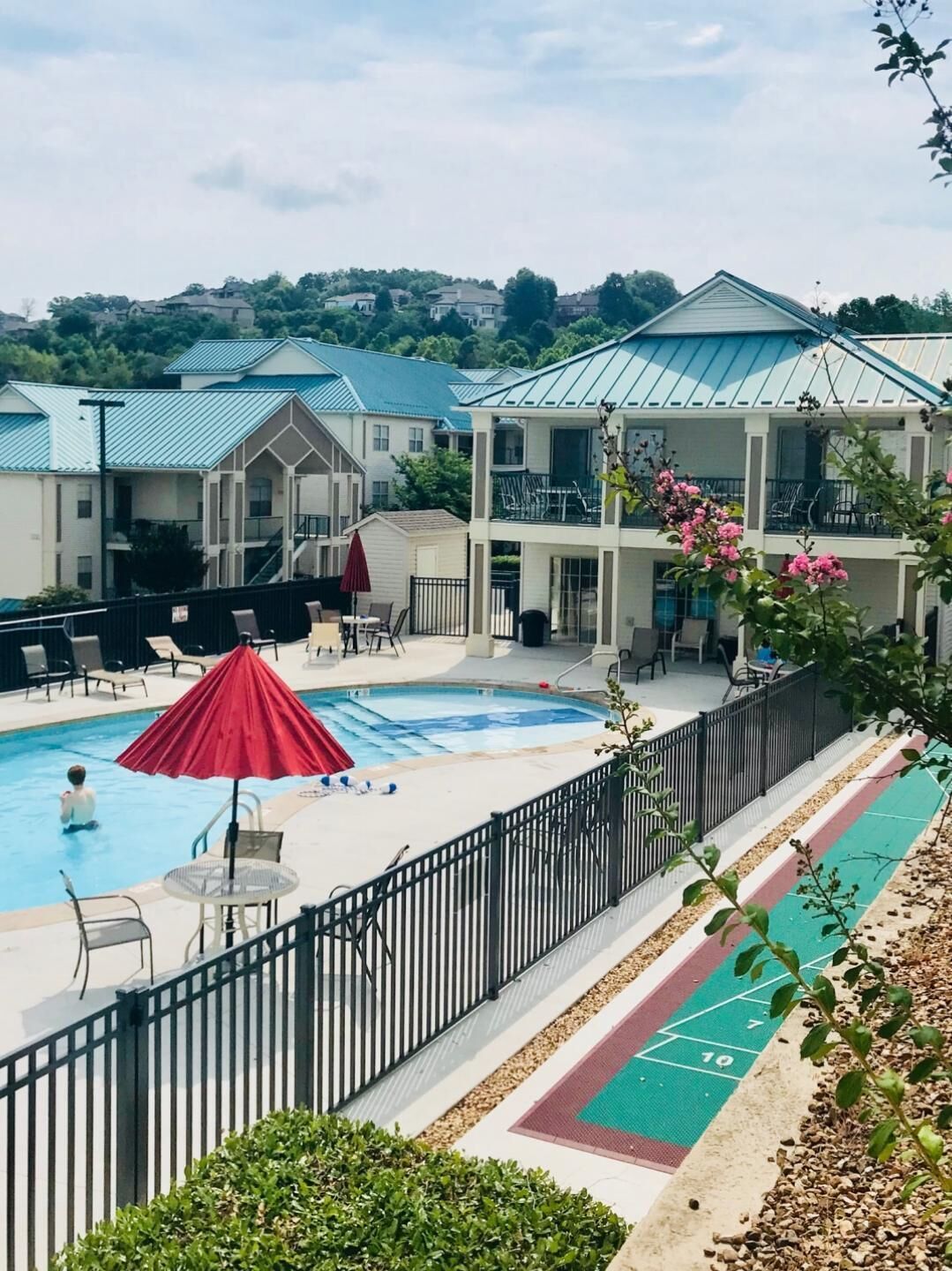 Ein Schwimmbecken mit Liegestühlen und Sonnenschirmen, umgeben von einem eingezäunten Bereich mit einem Basketballplatz.