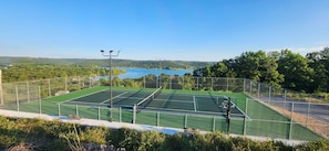 A tennis court with a net, surrounded by a fence, and a view of a lake and hills in the background.