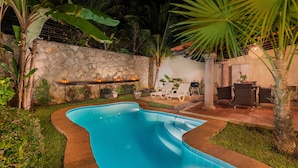 Una piscina con una parete in pietra e piante tropicali. Sono presenti anche lettini e un tavolo con sedie.