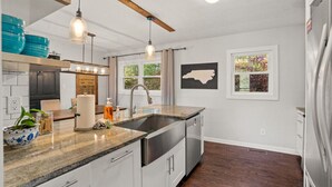 A modern kitchen with a granite countertop, stainless steel appliances, pendant lighting, and a window with a view of greenery.