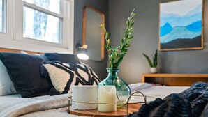A neatly arranged bedroom with a bed, a bedside table with a vase of greenery, a candle, and a cup.