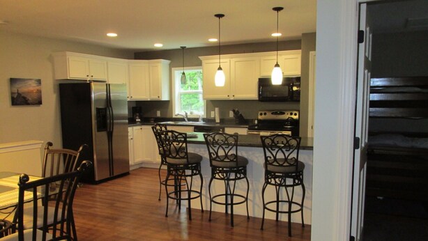 A modern kitchen with white cabinets, a stainless steel refrigerator, a microwave, and a stove. There are three bar stools and a dining table with chairs. - 2 Bedroom, Full Kitchen, Dining Room, Living Room, Large Deck, Jacuzzi -Sleeps 8 (Benzonia)