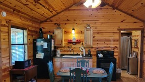 A cozy cabin-style kitchen with wooden walls, a dining table with chairs, a refrigerator, a stove, and a window with blinds.