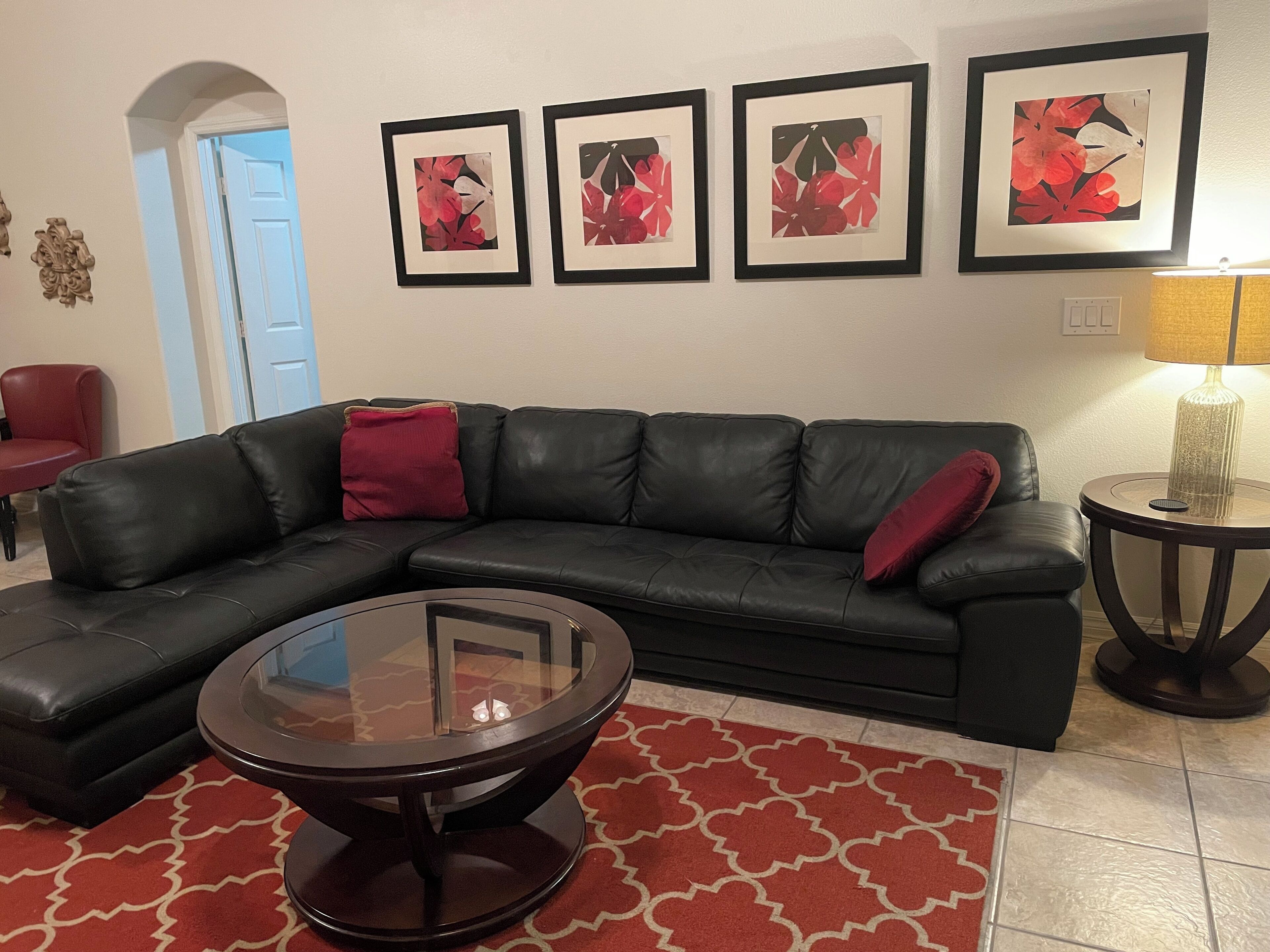A living room with a black leather sectional sofa, a glass coffee table, and a red patterned rug. There are three framed artworks on the wall and a lamp on a side table.