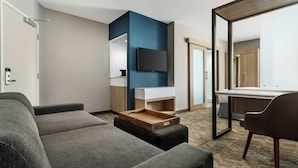 Une chambre d’hôtel moderne avec un canapé gris, une table d’appoint en bois, un téléviseur à écran plat fixé au mur et un grand miroir qui reflète la pièce.