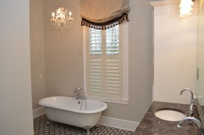 Une salle de bain avec une baignoire sur pieds, une fenêtre avec volets, un lustre et un lavabo en marbre.