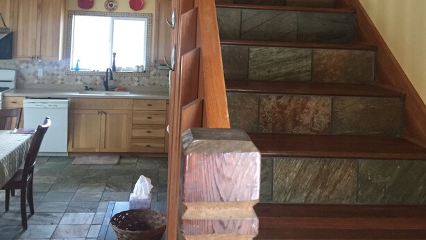 A wooden staircase with stone steps leading to a second floor. A dining area with a table and chairs is visible below. - Friendly Reeder Lake House (Kasilof)