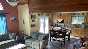 A cozy living room with a sofa, a wooden coffee table, and a dining table with chairs. A kitchen with wooden cabinets and a window is visible in the background. - Friendly Reeder Lake House (Kasilof)