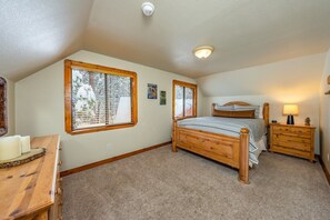 Ein Schlafzimmer mit einem hölzernen Bett, zwei Fenstern mit Jalousien, einem Nachttisch und einer Wandlampe.