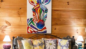 A cozy room with a wooden wall, a colorful zebra painting, a grey sofa with patterned cushions, and a small table with a lamp. - Climb On Inn ~Explore Red River Gorge & Comfort ~Perfect Location ~Pet Friendly  (Pine Ridge)