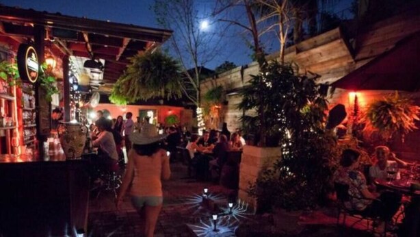 Un área de bar al aire libre con mesas y sillas, rodeada de plantas e iluminación decorativa.