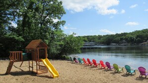 A sandy beach with a wooden play structure and a yellow slide, and colorful Adirondack chairs lined up along the water's edge. -  UPDATED LAKEFRONT WALK-IN UNIT, 2 POOLS, BEACH, DECK (Osage Beach)