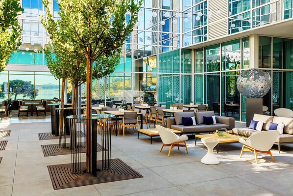 A modern outdoor seating area with tables and chairs, surrounded by glass walls and a large tree.