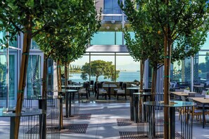 An outdoor dining area with tables and chairs, surrounded by glass walls and trees. - AC Hotel by Marriott San Francisco Airport Oyster Point/Waterfront (South San Francisco)