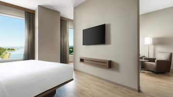 Una habitación de hotel moderna con una cama grande, un televisor de pantalla plana en la pared, una zona de estar con una silla y una mesita, y vistas al exterior a través de un ventanal amplio.