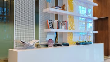 Un mostrador de recepción moderno con base blanca, múltiples estantes que exhiben libros y objetos decorativos, y una pared decorativa con un patrón geométrico.