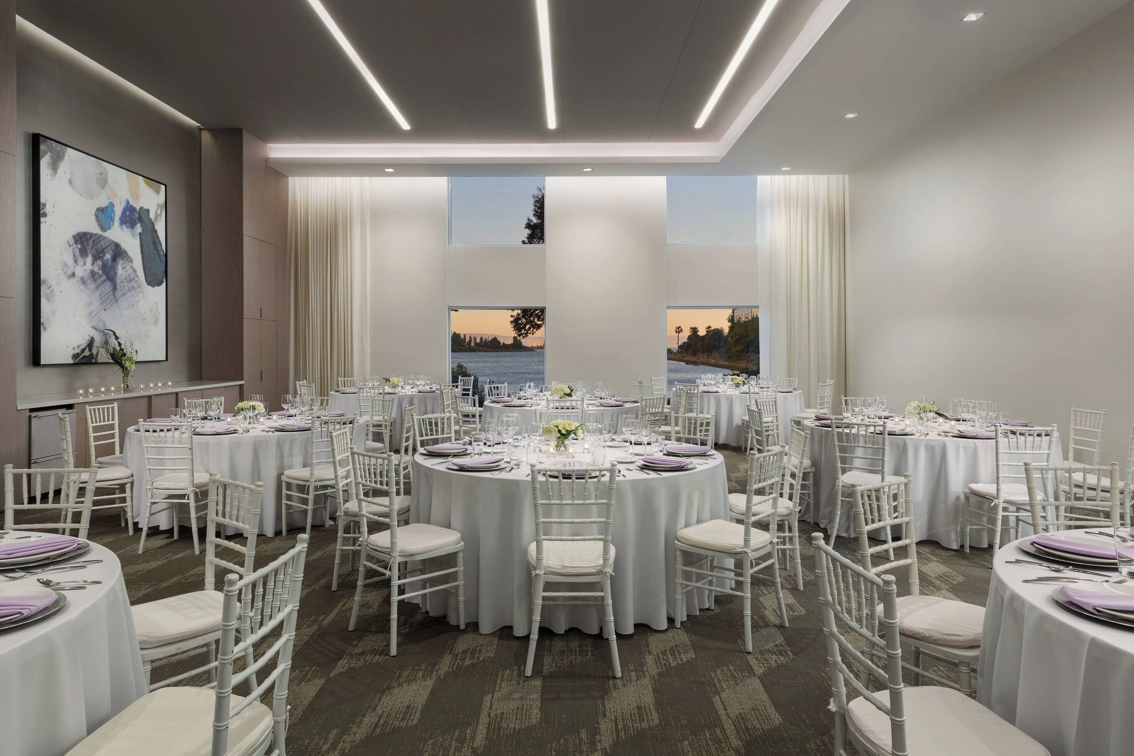 A banquet hall with round tables, white chairs, and a large painting on the wall.