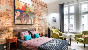 A bedroom with a brick wall, a bed with multiple pillows, a nightstand with a lamp, and a window with curtains. - Loft 1910 - Kazimierz Echoes (Kraków)