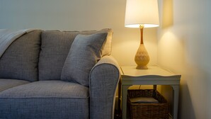 A grey sofa with a cushion, a small white side table with a lamp, and a wicker basket underneath.