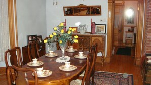 Une salle à manger avec une table en bois dressée pour quatre personnes, entourée de chaises. Un vase rempli de fleurs jaunes est posé sur la table, et une croix est accrochée au mur au-dessus.