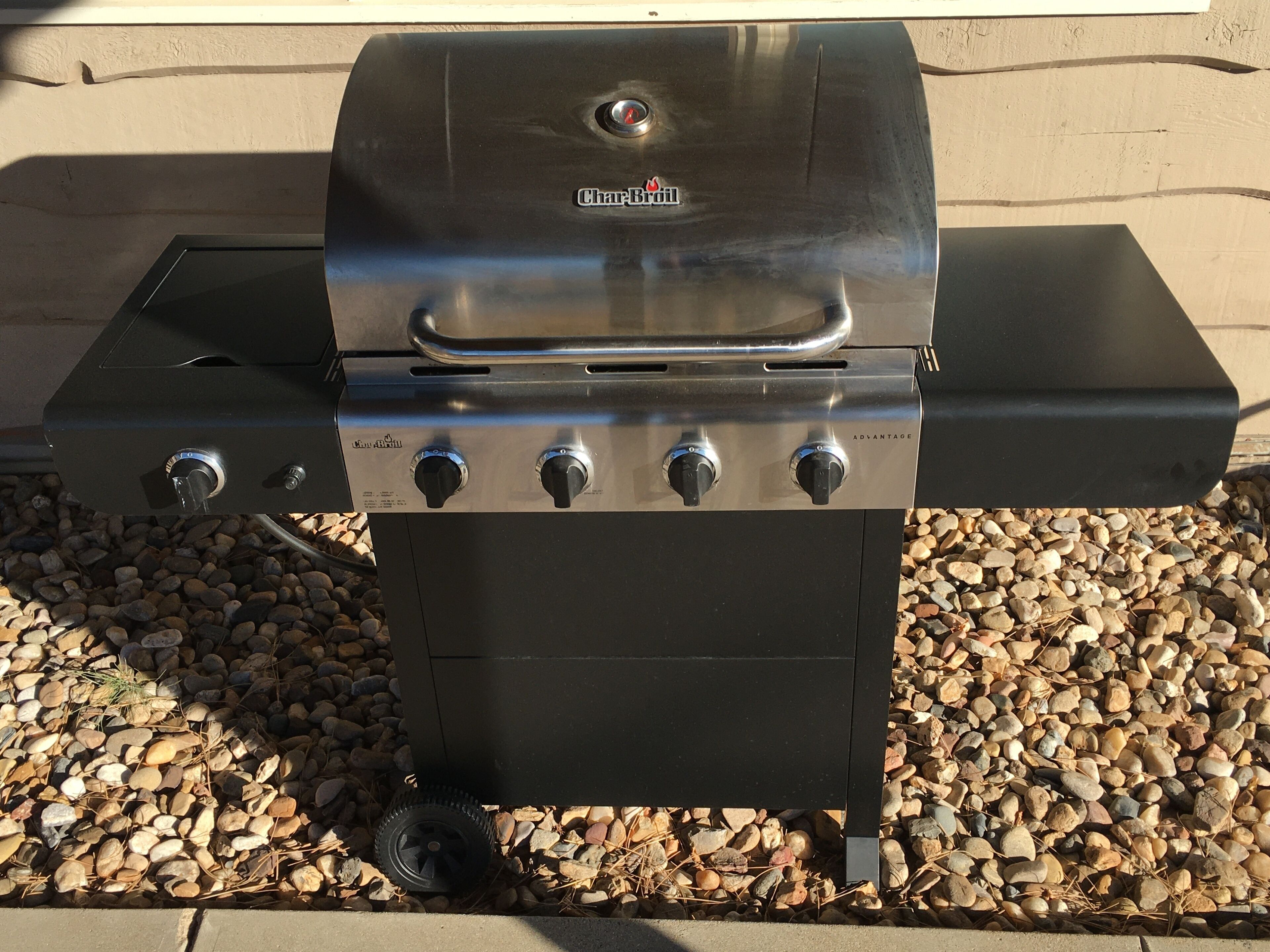 A Char-Broil gas grill with four burners and a wheel for mobility, placed on a gravel surface.