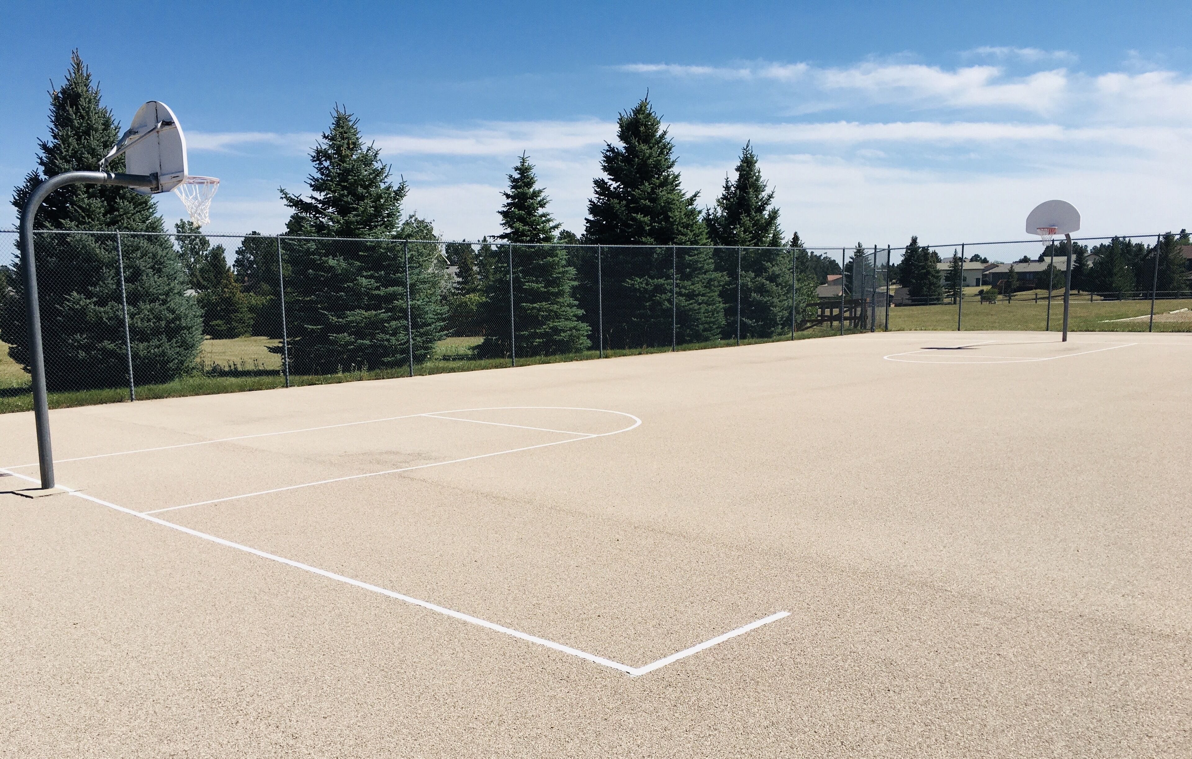 An outdoor basketball court with a hoop, surrounded by a chain-link fence and trees.