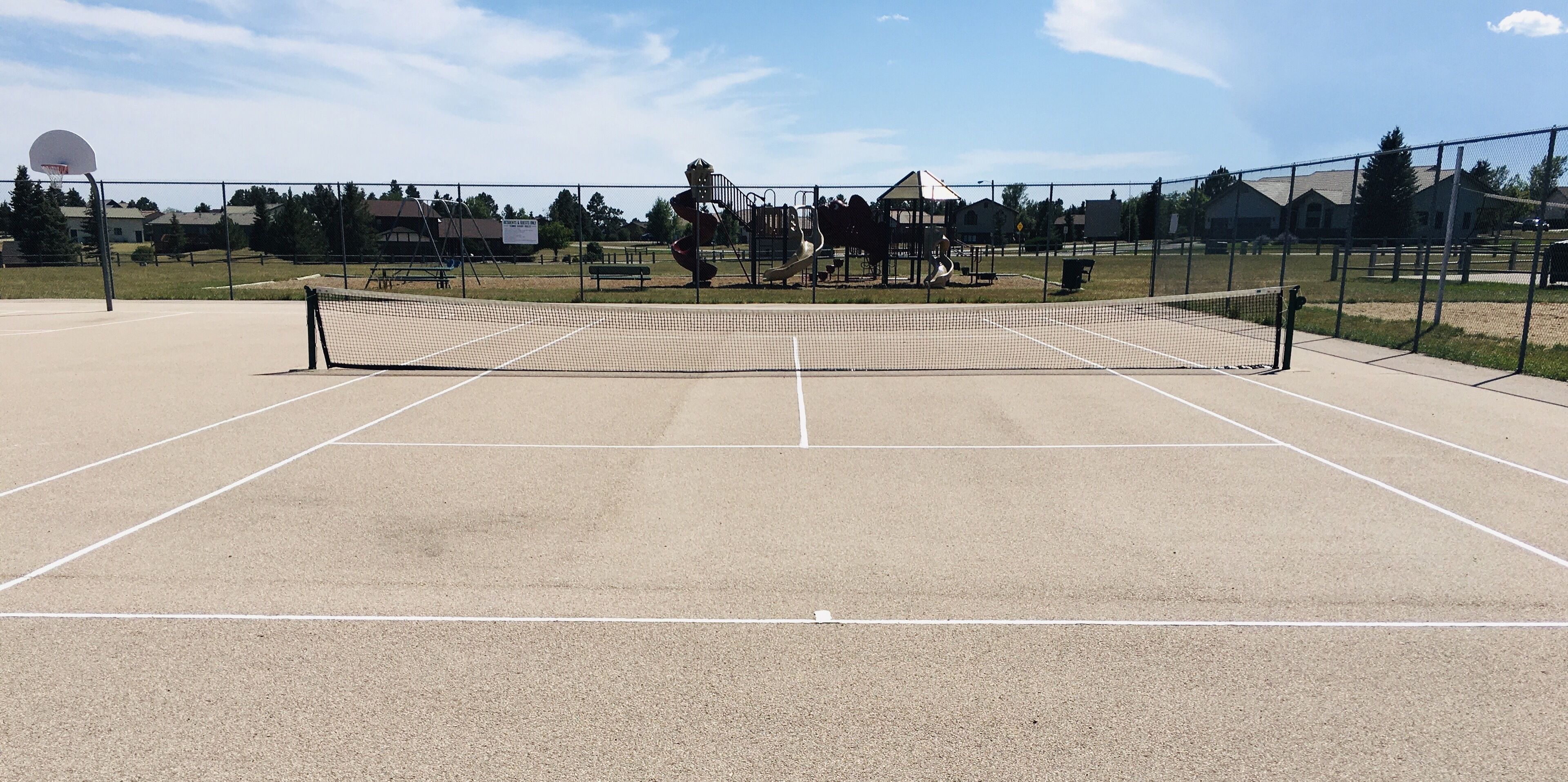 Una cancha de tenis exterior con red, rodeada por una cerca, y equipo de juegos infantiles al fondo.