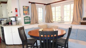 A dining area with a round table, four chairs, a window with curtains, and a kitchen area with cabinets and a fire extinguisher. - Perfect house for experiencing Mount Washington Valley year round! Pet Friendly! (North Conway)
