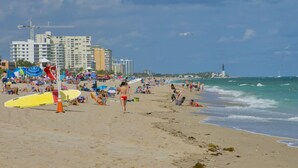 Uma praia com pessoas, guarda-sóis e um farol ao fundo.