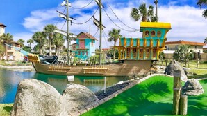 A colorful ship-shaped building with a green roof and yellow accents, situated next to a body of water with palm trees and a small dock. - Sol Mate-Elevator,Beach,Firepit rooftop,Arcade (St. Augustine)