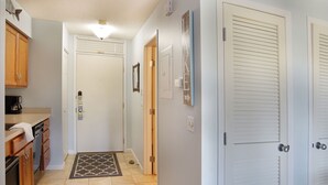 A hallway with a kitchen area on the left, a door with a mirror, and a wall-mounted light fixture. - 🏖️ Grand Traverse Bay Waterfront Condo • Beach, Sunsets, Park + More (Williamsburg)