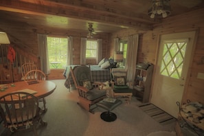 A cozy cabin interior with a dining table, chairs, a sofa, and a bed. There are windows allowing natural light and a door with a diamond-patterned glass.
