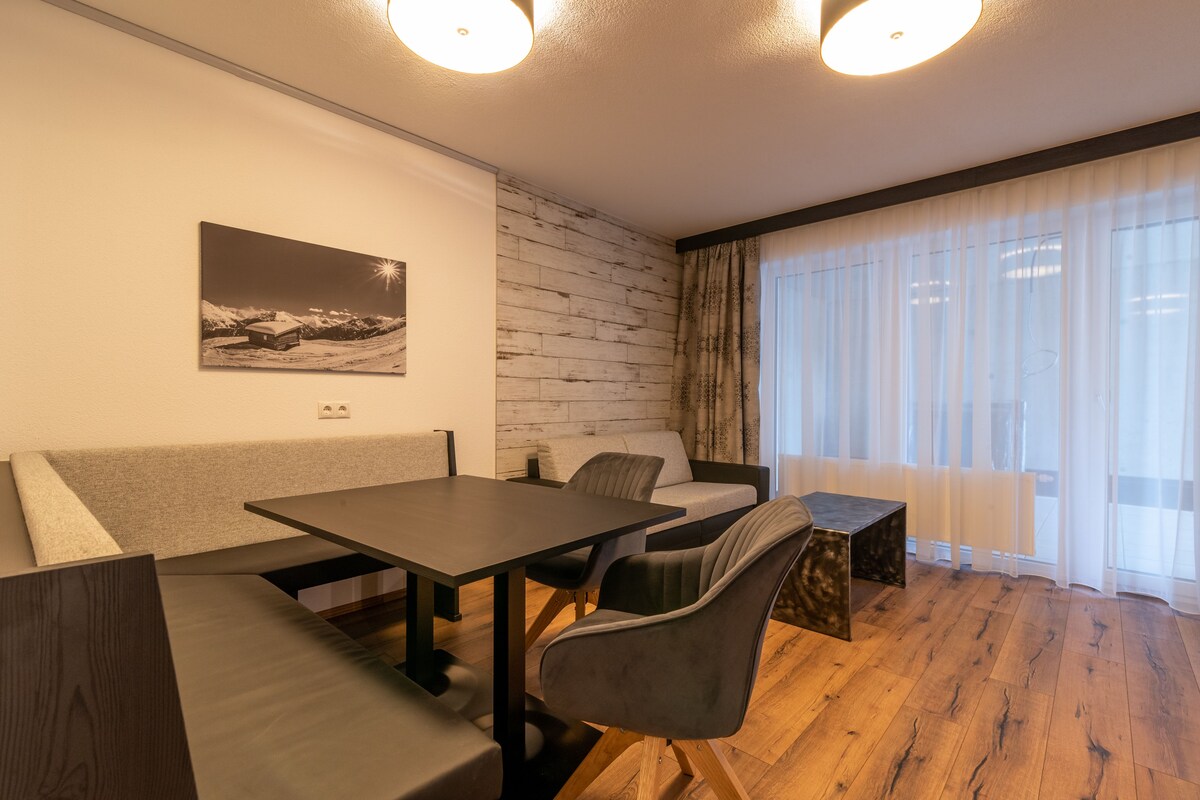 A modern room with a wooden floor, a dining table with chairs, a sofa, and a large window with sheer curtains.