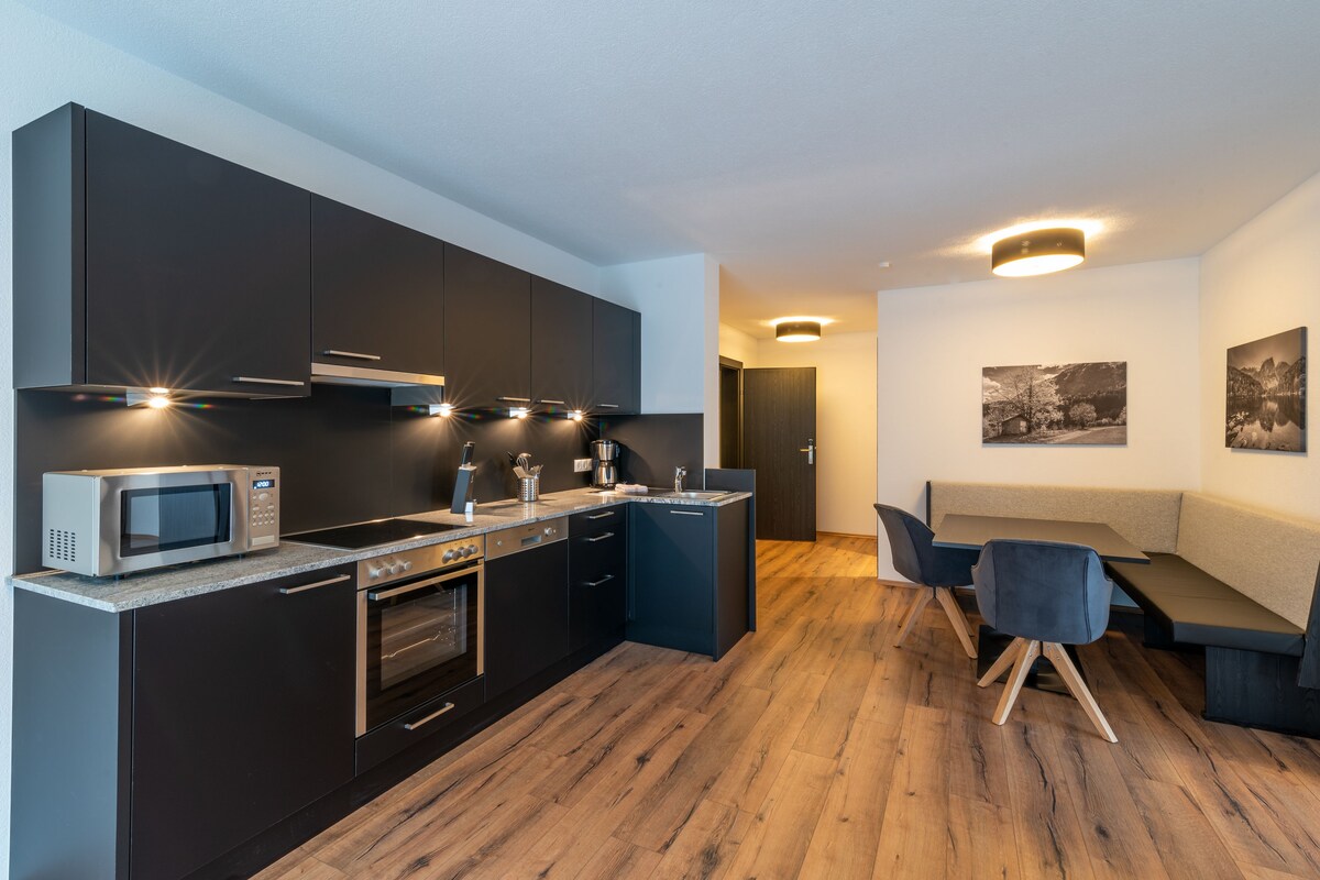 A modern kitchen with dark cabinetry, a built-in oven, and a microwave. There is a dining area with a bench and chairs.