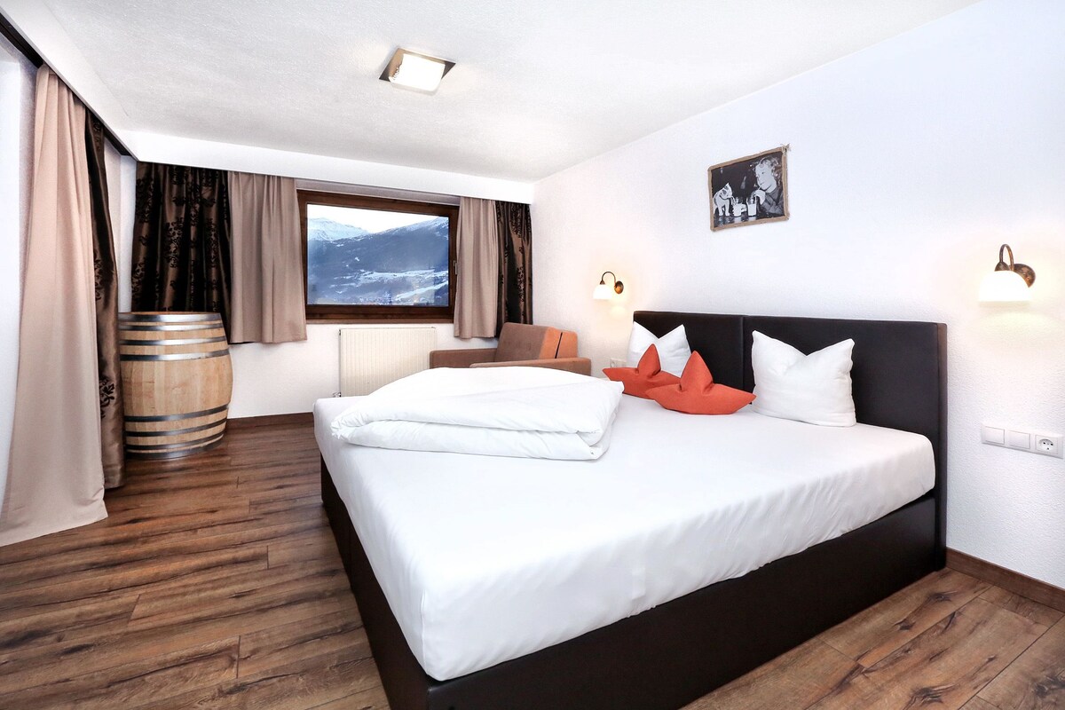 A hotel room with a large bed, wooden flooring, a window with a mountain view, a wooden barrel, and two wall-mounted lights.