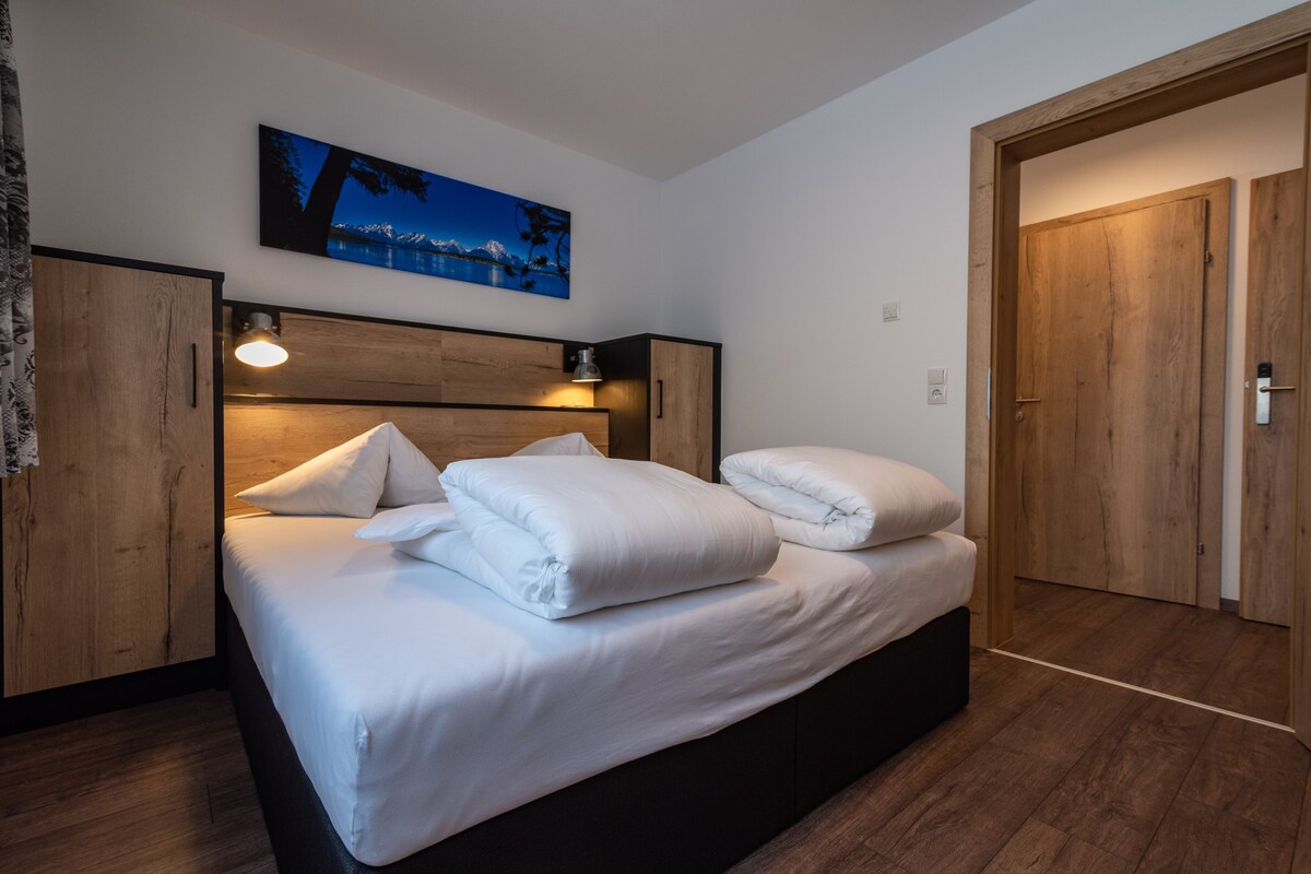 A modern bedroom with a large bed, wooden headboard, and a painting of a mountain landscape.