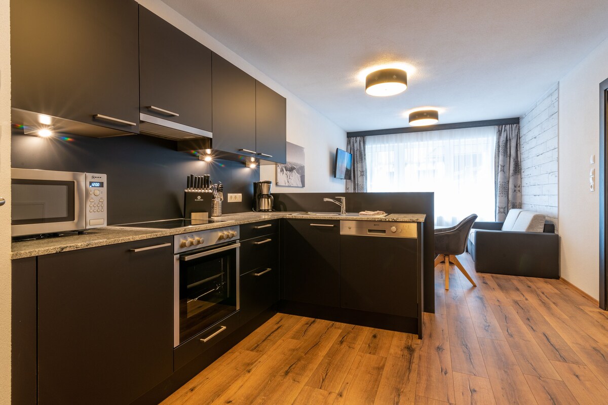 A modern kitchen with dark cabinetry, stainless steel appliances, and a wooden floor.
